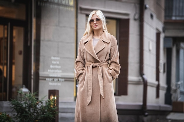 Jonge mooie blonde vrouw in een herfstjas staat op een straat in de stad. Zachte focus. Modieus begrip. Grijze achtergrond