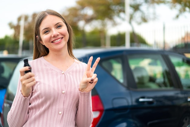 Jonge mooie blonde vrouw met autosleutels buitenshuis glimlachend en overwinningsteken tonen