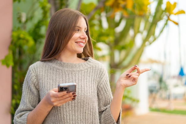 Jonge mooie blonde vrouw met behulp van mobiele telefoon buitenshuis wijzend naar de zijkant om een product te presenteren