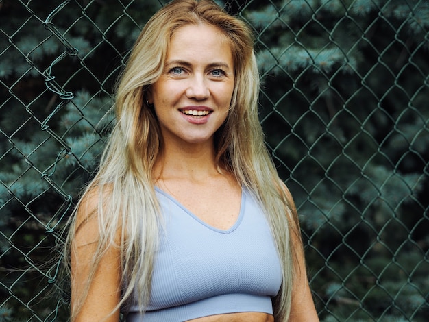 Foto jonge, mooie blonde vrouw met lang haar in lichtblauwe activewear glimlachend met gaas op de achtergrond