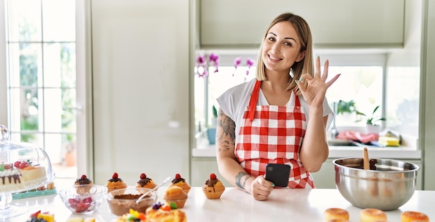 Jonge mooie blonde vrouw met schort kokende gebakjes op zoek naar recept op smartphone die ok teken doet met vingers glimlachend vriendelijk gebarend uitstekend symbool