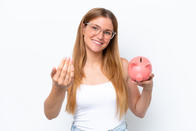 Jonge mooie blonde vrouw met spaarvarken geïsoleerd op een witte achtergrond die uitnodigt om met de hand te komen Blij dat je kwam