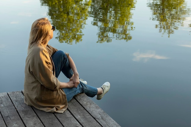 Jonge mooie blonde vrouw zit op een houten kade en geniet van het uitzicht op het meer in de zomer tijdens de zonsondergang en rust