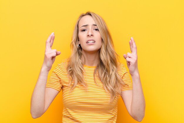 Jonge mooie blondevrouw die angstig vingers kruisen en op geluk met een ongerust gemaakte blik tegen vlakke kleurenmuur hopen