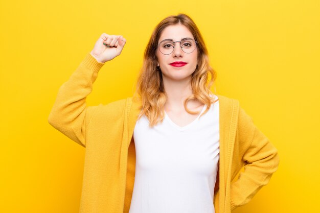 Jonge mooie blondevrouw die ernstig, sterk en opstandig voelen, vuist opsteken, protesteren of vechten voor revolutie tegen gele muur