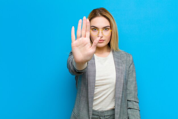 Jonge mooie blondevrouw die ernstig, streng, ontevreden en boos tonend open palm kijkend eindegebaar maken tegen vlakke muur