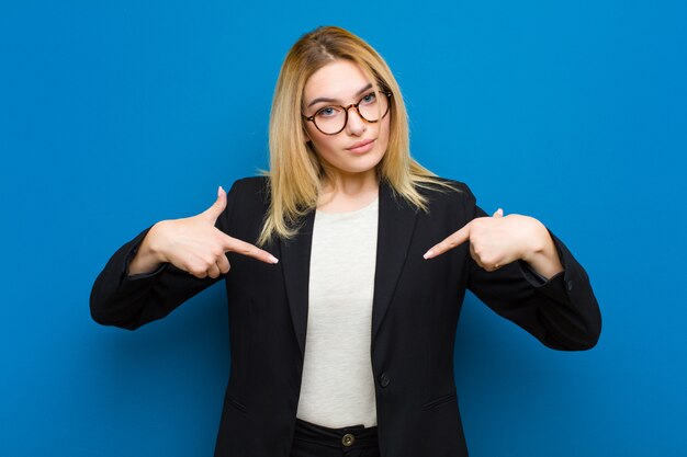 Jonge mooie blondevrouw die trots, positief en toevallig het richten aan borst met beide handen tegen vlakke muur kijken