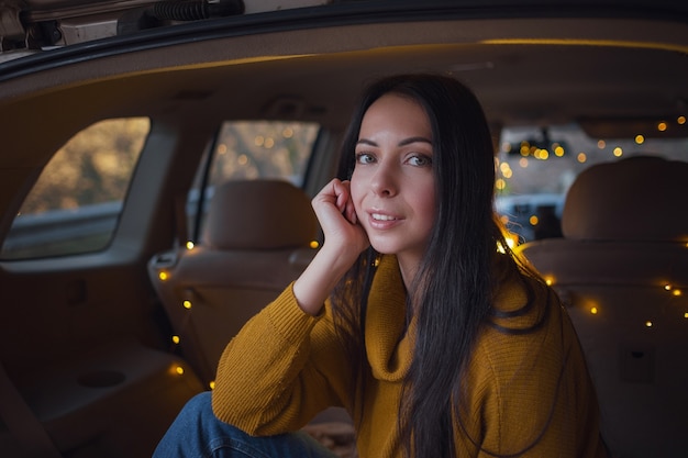 Jonge mooie brunette geniet van ontspannen in de kofferbak van haar auto. een gele slinger en een beige plaid zorgen voor gezelligheid