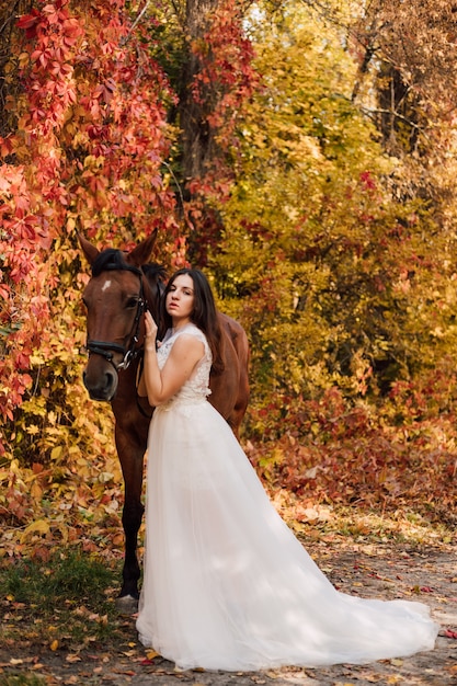 Jonge mooie brunette in een witte jurk knuffelt een bruin paard