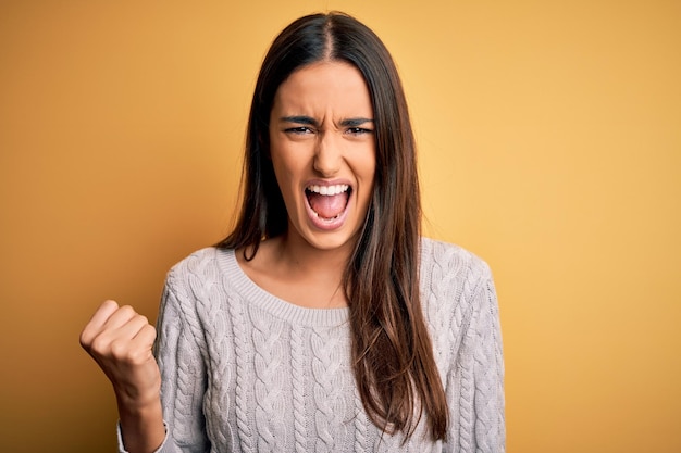 Jonge mooie brunette vrouw, gekleed in een witte casual trui over een gele achtergrond, boos en gek, vuist gefrustreerd en woedend opstekend terwijl ze schreeuwde van woede Woede en agressief concept