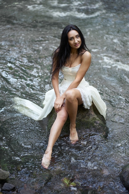 Jonge mooie brunette vrouw in witte trouwjurk zittend op de rots in wervelende rivier