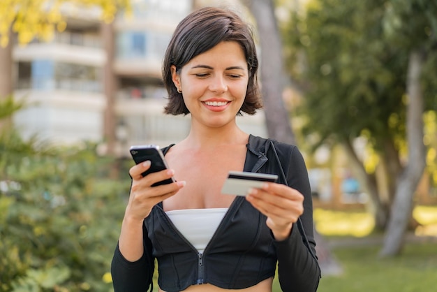 Jonge mooie Bulgaarse vrouw die buitenshuis met de mobiel koopt met een creditcard