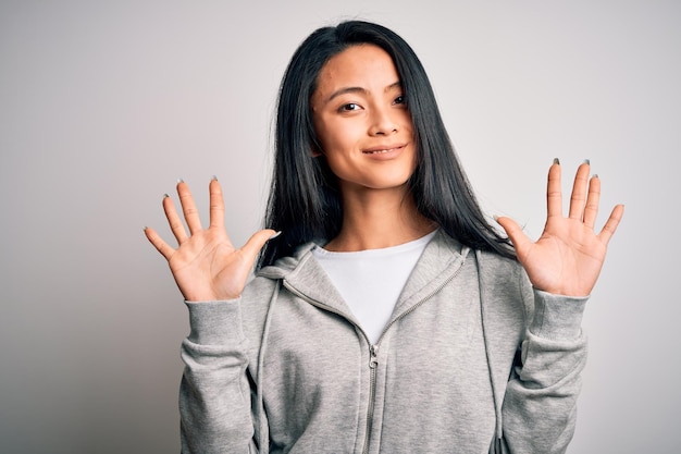 Jonge mooie chinese sportieve vrouw die sweatshirt over geïsoleerde witte achtergrond draagt en met vingers nummer tien naar boven wijst terwijl ze zelfverzekerd en gelukkig glimlacht