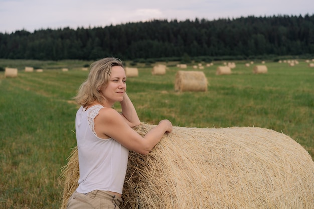 Jonge mooie dame met blond haar leunt op hooiberg en kijkt opzij in het zomerveld