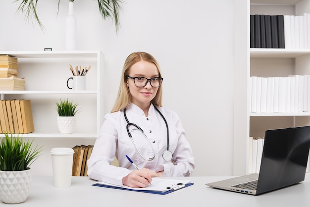 Jonge mooie dokter vrouw gelukkig werken en glimlach in het ziekenhuis, zittend op tafel, medische concept