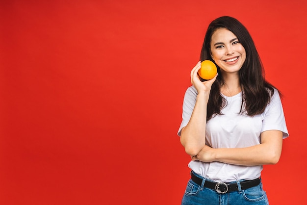 Jonge mooie donkerbruine vrouw die oranje fruit houdt dat over rode achtergrond wordt geïsoleerd