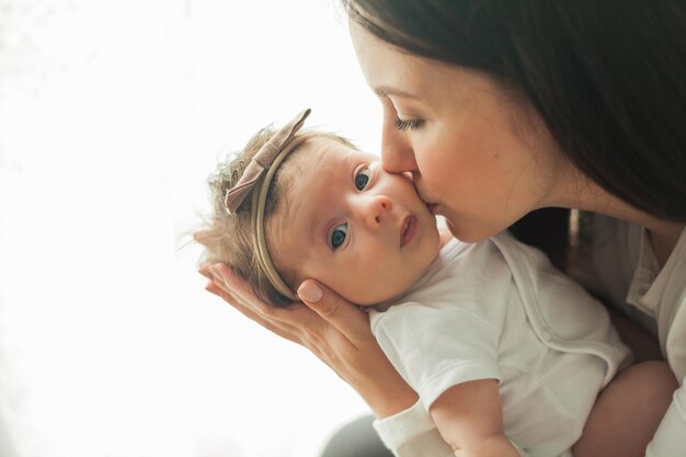 Jonge mooie donkerharige moeder houdt haar pasgeboren dochter in haar armen in een gezellig huis Familieportret Moederschap Jonge vrouw kust haar 2 maanden oude baby