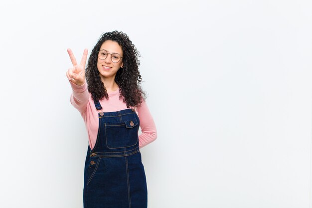 Jonge mooie en vrouw die gelukkig, onbezorgd en positief, gesturing overwinning of vrede met één hand glimlachen kijken tegen witte muur