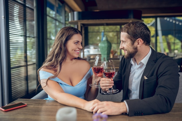 Foto jonge mooie geliefden man en vrouw in mooie kleren zitten met glazen wijn aan tafel in café