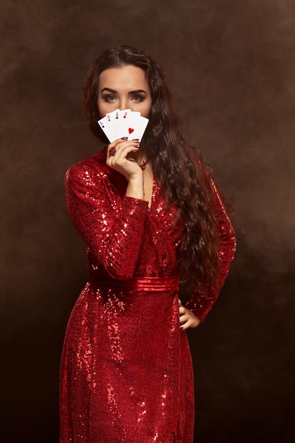 Jonge mooie gelukkige brunette in een rode avondjurk toont haar kaarten een vier van azen die haar gezicht verbergen...
