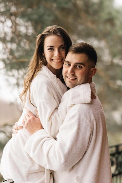 Jonge mooie gelukkige pasgetrouwden verliefd een man en een vrouw in witte badjassen op het balkon van het huis glimlachen en knuffelen