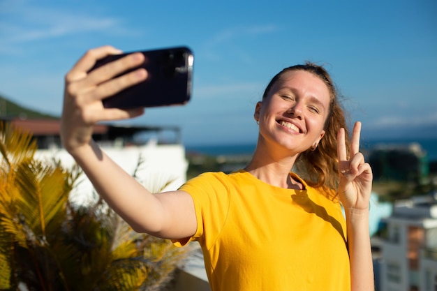 Jonge mooie gelukkige vrouw blogger neemt selfie foto van zichzelf op frontale camera naar sociale media van smartphone mobiele telefoon glimlachend zwaaiend met haar hand met video-oproepgesprek