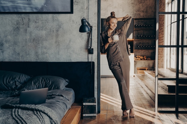 Foto jonge mooie gelukkige vrouw die een bruine satijnen pyjama draagt, een koffiekopje vasthoudt terwijl ze in de deuropening van de slaapkamer staat nadat ze 's ochtends thuis wakker is geworden, vrouw die een nieuwe dag begint