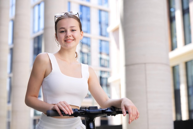 Jonge mooie gelukkige vrouw vrij aantrekkelijk meisje tiener rijdt electro scooter in de stad op zonnige zomerdag in witte kleren glimlachend in zonnebril elektrische eco delen vervoer