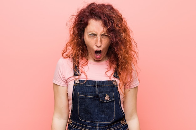 Jonge mooie gember roodharige vrouw die een jeans dungaree draagt die erg boos en agressief gilt.