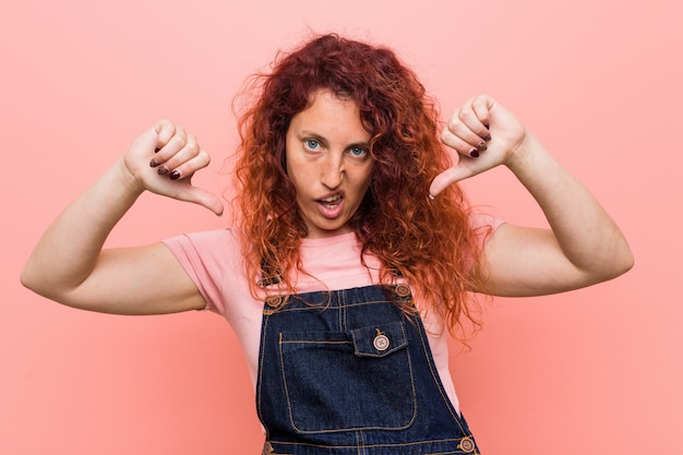 Jonge mooie gember roodharige vrouw draagt een denim tuinbroek tonen duim naar beneden en uiten van afkeer