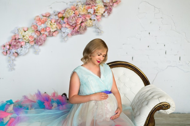Jonge mooie glamour zwangere vrouw in een kleurrijke gezwollen jurk staat in een bloemenzolder. Gelukkig zwangerschap