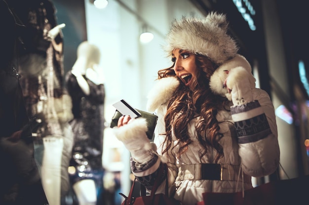 Jonge, mooie glimlachende vrouw die plezier heeft in de straat van de stad met Kerstmis en een foto maakt met smartphone door de etalagewinkel.