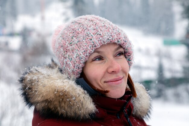 Jonge mooie glimlachende vrouw die zijdelings in roze gebreide hoed met pompon in warm jasje in openlucht kijkt