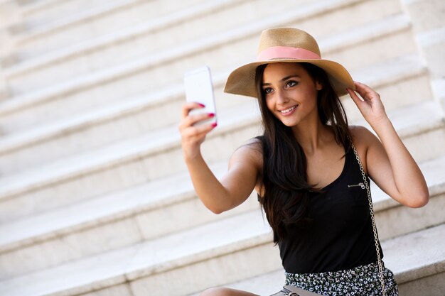 Jonge mooie glimlachende vrouw in hoed die in openlucht selfie met smartphone maken