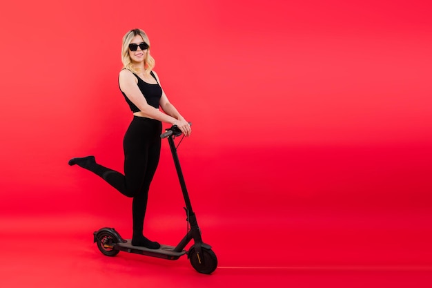 Jonge, mooie glimlachende vrouw in sportkleding die met een elektrische scooter staat, kijkt gelukkig in de camera