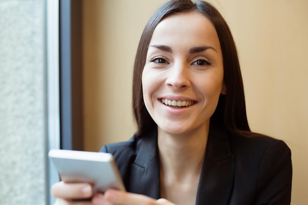 Jonge mooie glimlachende zakenvrouw in een zwarte jas die aan de telefoon werkt of internet gebruikt terwijl ze aan een tafel in een café zit