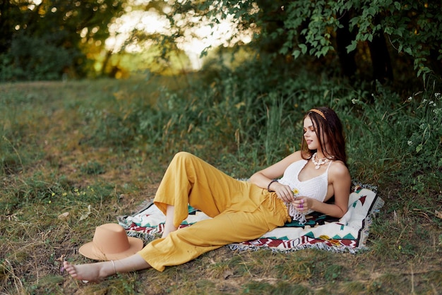 Jonge mooie hippievrouw liggend op de grond in de natuur in de herfst in ecokleding in gele broek in het zonsonderganglicht