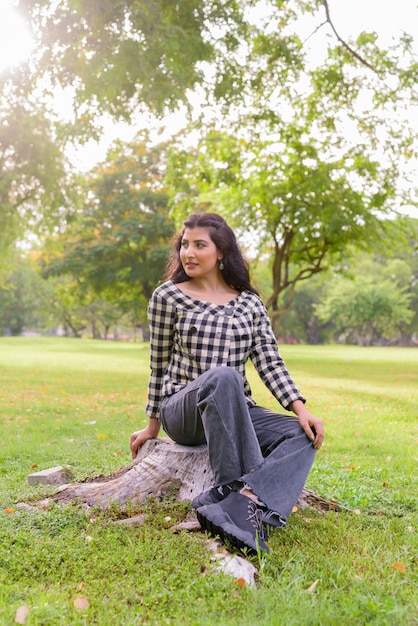 Jonge mooie Indiase hipster vrouw denken en zitten in het park buiten