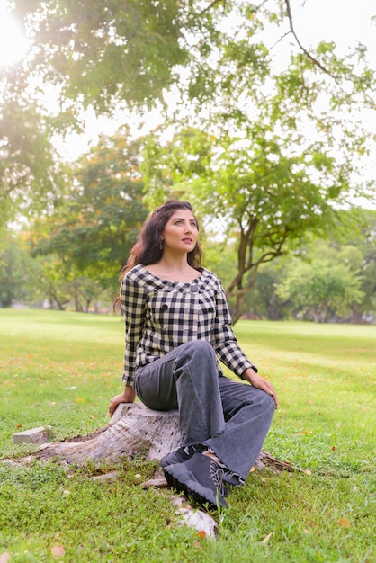 Jonge mooie Indiase hipster vrouw denken en zitten in het park buiten