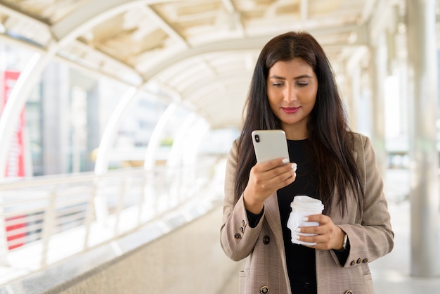 Jonge mooie Indiase zakenvrouw met behulp van telefoon en koffie onderweg in de stad