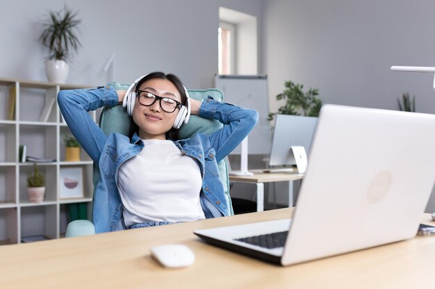 Jonge, mooie kantoormedewerker die ontspant aan het bureau aziatische vrouw die tijdens werkpauze naar muziek luistert in een koptelefoon