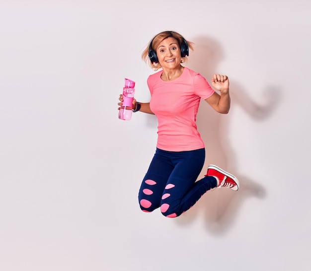 Jonge mooie kaukasische sportvrouw die sportkleding draagt die gelukkig glimlacht Springend met een glimlach op het gezicht met een fles water over een geïsoleerde witte achtergrond