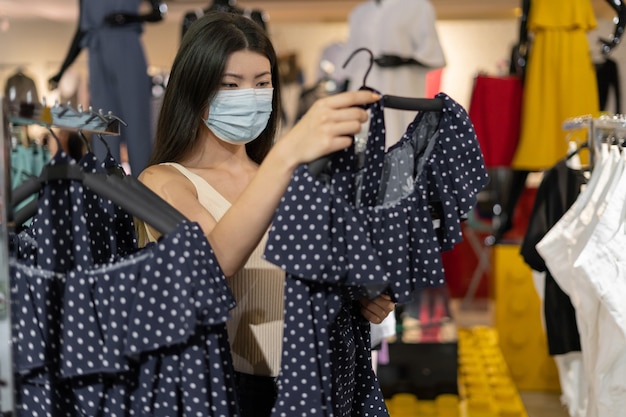 Jonge mooie koreaanse vrouw gekleed in een beschermend medisch masker op haar gezicht staat in een kledingwinkel. dingen kopen tijdens een pandemie.