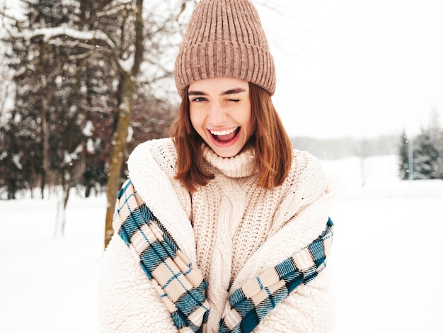 Jonge mooie lachende hipster vrouw in trendy warme kleding en sjaal. Zorgeloos vrouw poseren in de straat in het park. Positief puur model met plezier in de sneeuw. Genieten van winterse momenten. Knipogen