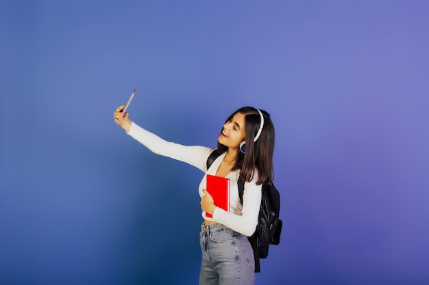 Jonge mooie lachende student meisje met zwarte rugzak en koptelefoon maakt selfie op haar telefoon op blauwe ondergrond.