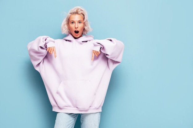 Foto jonge mooie lachende vrouw in trendy zomer hoodie sexy zorgeloze vrouw poseren in de buurt van blauwe muur in studio positieve blond model plezier en gek te wijzen op gratis mockup ruimte