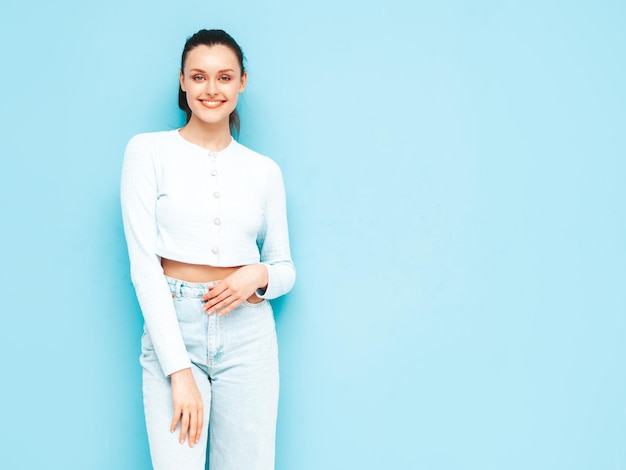 Jonge mooie lachende vrouw in trendy zomer jeans en topkleren sexy zorgeloze vrouw poseren in de buurt van blauwe muur in studio positieve brunette model plezier vrolijk en gelukkig