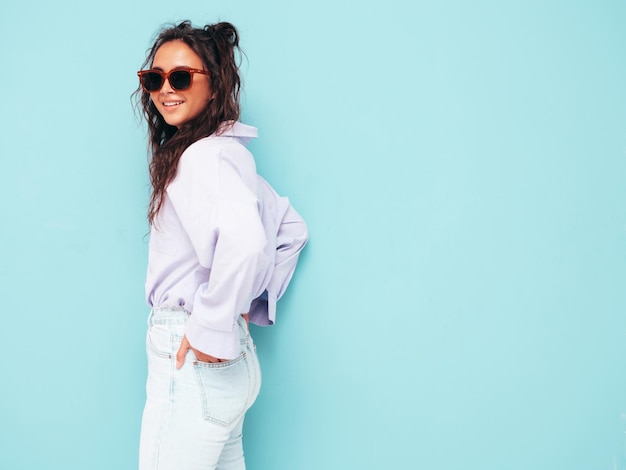 Jonge mooie lachende vrouw in trendy zomerkleren Sexy zorgeloze vrouw poseren in de buurt van blauwe muur in studio Positieve brunette model plezier en gek vrolijk en gelukkig in zonnebril