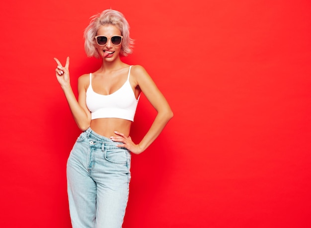 Jonge mooie lachende vrouw in trendy zomerkleren Sexy zorgeloze vrouw poseren in de buurt van rode muur in studio Positieve blond model plezier en gek vrolijk en gelukkig In zonnebril