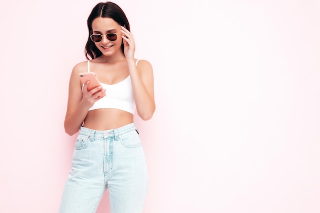 Jonge mooie lachende vrouw in trendy zomerkleren Sexy zorgeloze vrouw poseren in de buurt van roze muur in studio Positieve brunette model kijken naar mobiele telefoon scherm Holding smartphone en het gebruik van apps